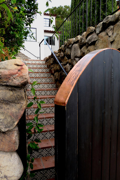 a Beautiful Dark Stained Spanish Entry Gate Made of Wood Planks and with a Copper Cap is in the Foreground of a Spanish Tile Staircase Leading Up to a Spanish House in the Riviera Neighborhood of Santa Barbara, California