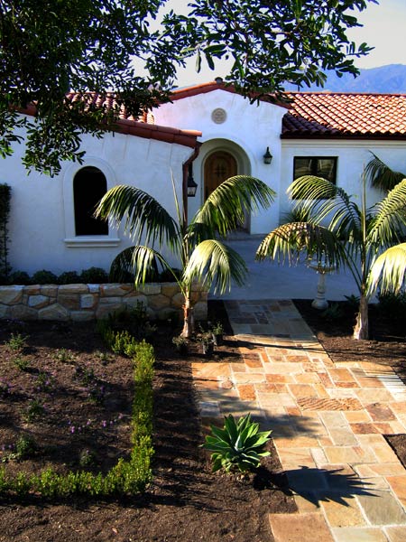 A top ten best Spanish style house design by Jeff Doubét includes a small entry tower with round top wood front door, as well as round top windows in the garage that have special plaster surround and window sill details. Custom built local Santa Barbara sandstone walls for planters, as well as a flagstone walkway from the street was installed.