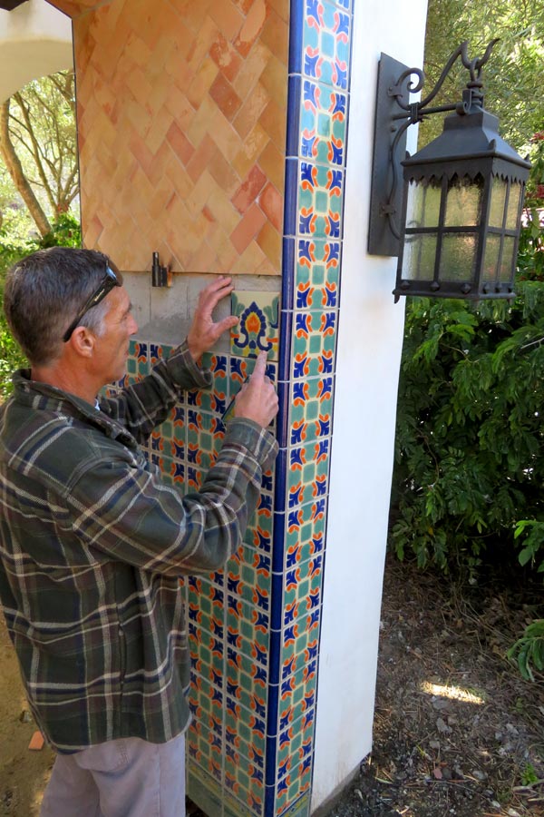 A talented tile installer installs decorative spanish tiles on an outdoor column. Part of a charming spanish archway leading to a santa barbara garden, beyond.