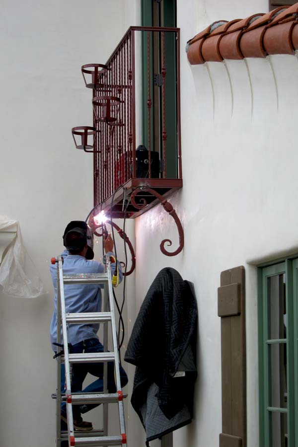 A welder stands on a ladder while welding decorative iron support brackets to a spanish juliette balcony that is being installed on a two story spanish home in santa barbara, ca